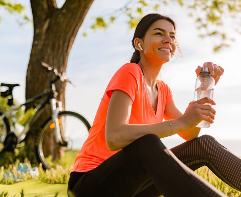 smiling beautiful woman drinking water in bottle doing sports in morning in park nature doing yoga on mat, pink fitness outfit, happy healthy lifestyle, music in earphones, bicycle on background